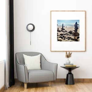 May include: A grey armchair with a white cushion sits next to a black side table with a vase of dried flowers and books. A framed photograph of a beach scene with stacked rocks hangs on the wall above the table. A round, white, wall-mounted light fixture is visible in the top left corner of the image.