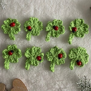 Könnte beinhalten: Acht gehäkelte grüne vierblättrige Kleeblätter mit roten Marienkäfern auf einer weißen flauschigen Oberfläche.