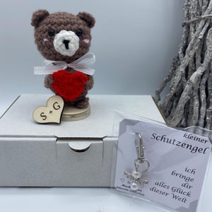 Könnte beinhalten: Ein brauner gehäkelter Teddybär, der ein rotes Herz hält. Der Bär sitzt auf einem Holzsockel mit einem Holzhäuschen, auf dem "S+G" steht. Der Bär befindet sich in einer weißen Box mit einer kleinen weißen Geschenktüte, die einen silbernen Anhänger mit einer Perle und Flügeln enthält. Der Anhänger hat ein Etikett mit der Aufschrift "kleiner Schutzengel ich bringe dir alles Glück dieser Welt".