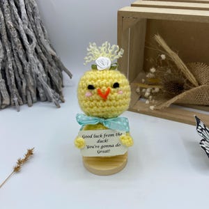 Könnte beinhalten: Handgefertigte gelbe gehäkelte Entenfigur mit hellblauer Schleife und einem kleinen Schild mit der Aufschrift "Good luck from the duck! You're gonna do Great!". Die Ente hat eine weiße Blume und gelbe Akzente.