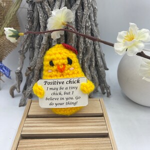 Könnte beinhalten: Ein kleines gelbes Häkelküken, das ein Schild mit der Aufschrift "Positive chick I may be a tiny chick, but I believe in you. Go do your thing!" hält.