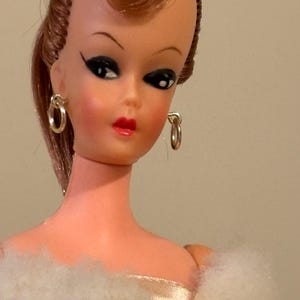 May include: A vintage doll with auburn hair styled in a classic updo, wearing gold hoop earrings. The doll has a fair complexion, dark eye makeup, and red lipstick. She is dressed in a white, fluffy, faux fur stole over a satin dress.