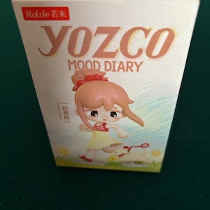 Puede incluir: Una caja de cartón blanca rectangular con el logotipo de la marca Rolife, el texto YOZCO MOOD DIARY y una ilustración de un personaje con cabello castaño claro en coleta, vistiendo una blusa a cuadros rojos y blancos y una falda amarilla.