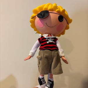 Pode incluir: Boneca Lalaloopsy vestida de pirata. A boneca tem cabelo amarelo, tapa-olho preto e um olho de botão. Ela usa uma camisa listrada vermelha e preta com uma caveira, shorts de veludo cotelê e tênis preto e branco. A boneca está em um suporte branco.
