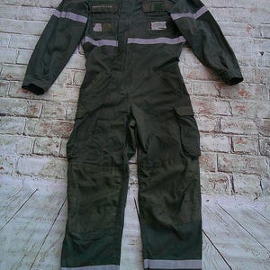 Puede incluir: Un traje de vuelo verde oliva con cuello alto, mangas largas y múltiples bolsillos en el pecho y las piernas. La prenda incluye bandas reflectantes plateadas horizontales en el pecho, brazos y piernas, con el texto ARMÉE DE L'AIR impreso.