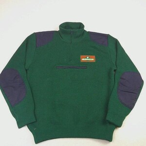 May include: A dark green quarter-zip sweater featuring navy blue shoulder patches and elbow patches. The garment has a high collar and a rectangular patch on the left chest with the text The Golfing Life in white and green lettering.