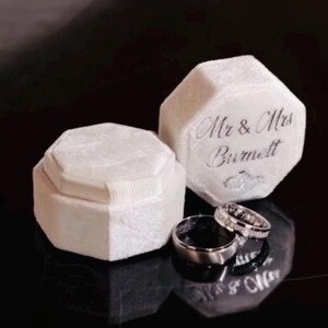 May include: Two white velvet octagonal ring boxes with a textured finish. One box is closed, while the other is open and features the silver script text 'Mr & Mrs Burnett' on the lid. Two silver-toned metal rings rest in front of the boxes.
