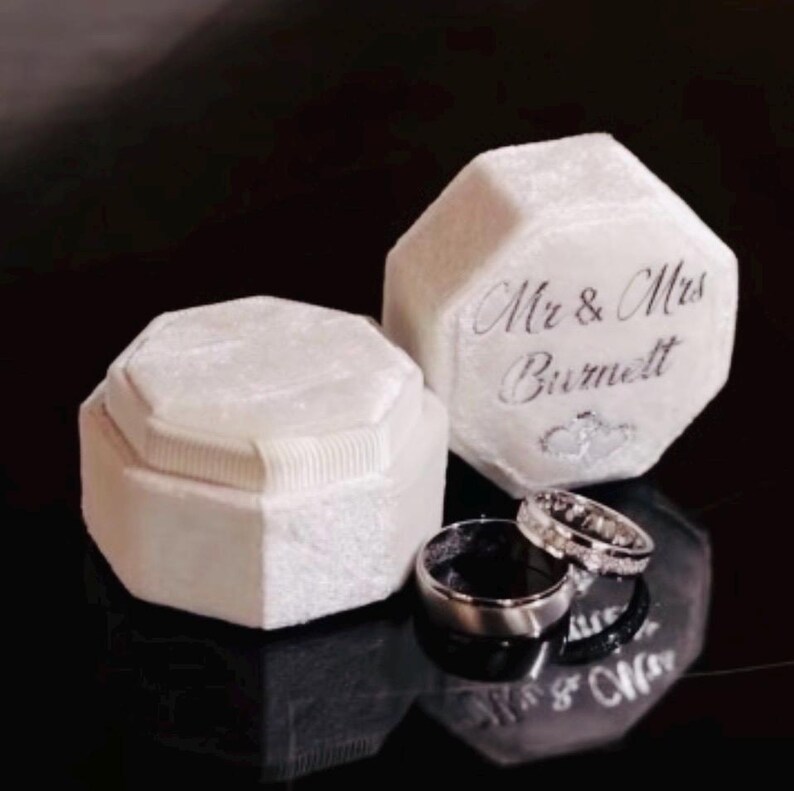 May include: Two white velvet octagonal ring boxes with a textured finish. One box is closed, while the other is open and features the silver script text 'Mr & Mrs Burnett' on the lid. Two silver-toned metal rings rest in front of the boxes.