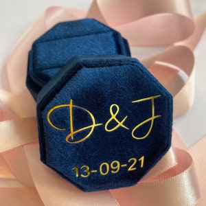 May include: A navy blue velvet octagonal ring box featuring gold lettering that reads D & J and the date 13-09-21. The box has a hinged lid and a soft, textured fabric finish, designed to hold jewelry securely within its interior compartment.