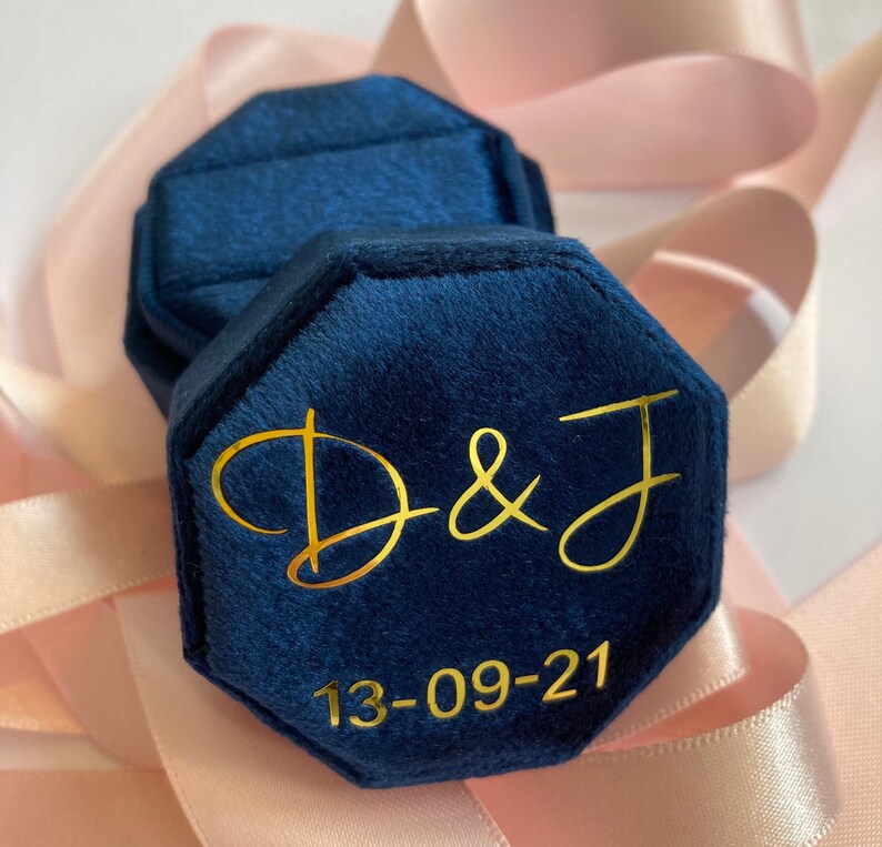 May include: A navy blue velvet octagonal ring box featuring gold lettering that reads D & J and the date 13-09-21. The box has a hinged lid and a soft, textured fabric finish, designed to hold jewelry securely within its interior compartment.