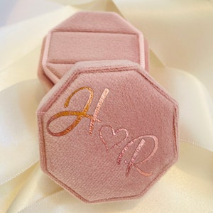 May include: An octagonal dusty pink velvet ring box with a hinged lid. The top of the lid features the metallic copper-colored script letters H and R separated by a small heart symbol. The box has a soft, fuzzy texture and a structured shape.