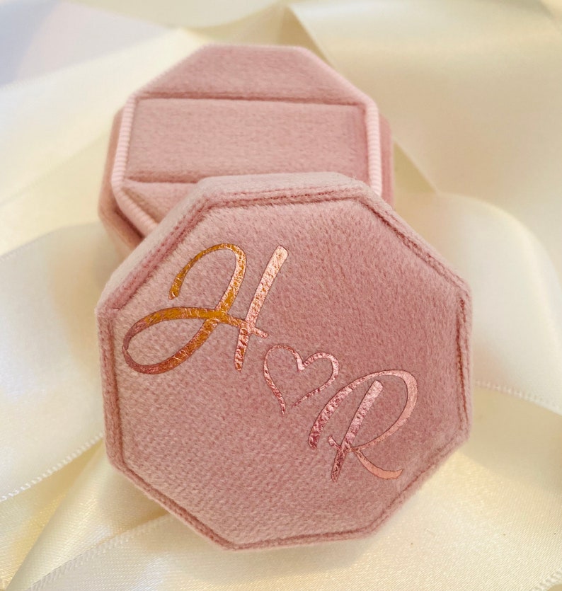 May include: An octagonal dusty pink velvet ring box with a hinged lid. The top of the lid features the metallic copper-colored script letters H and R separated by a small heart symbol. The box has a soft, fuzzy texture and a structured shape.