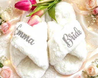 May include: A pair of white faux fur slippers with a crisscross strap design. The left slipper features the name Emma in silver glitter script, and the right slipper features the word Bride in silver glitter script. The slippers are plush and soft.