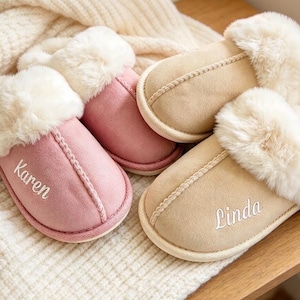 May include: Two pairs of slip-on slippers with white faux fur trim. One pair is dusty pink with the name Karen embroidered in white, and the other pair is beige with the name Linda embroidered in white. Both feature a suede-like exterior finish.