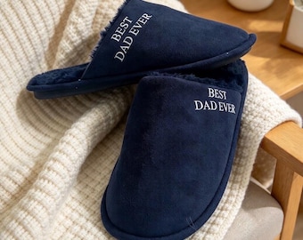 May include: A pair of navy blue slippers with a soft, dark interior lining. The top of each slipper features the white embroidered text "BEST DAD EVER" in a clean, sans-serif font. The slippers have a smooth, matte exterior finish.