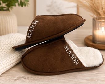May include: A pair of brown suede slippers with white faux fur lining. The exterior of each slipper features the name MASON embroidered in white capital letters. The slippers have a dark sole and a slip-on design with an open back construction.