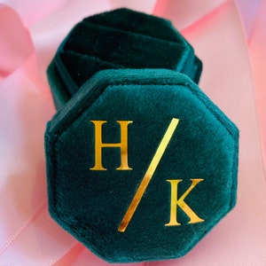 May include: An octagonal dark green velvet ring box featuring a gold foil monogram with the letters H and K separated by a diagonal slash. The lid is removable, revealing a matching dark green velvet interior designed to hold a single ring.