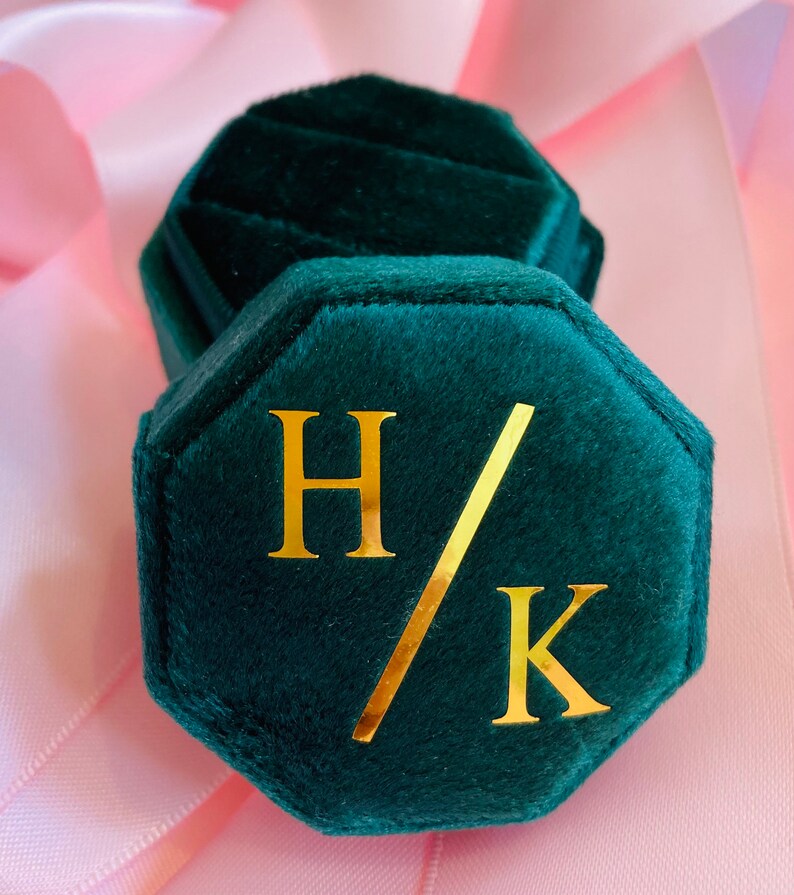 May include: An octagonal dark green velvet ring box featuring a gold foil monogram with the letters H and K separated by a diagonal slash. The lid is removable, revealing a matching dark green velvet interior designed to hold a single ring.