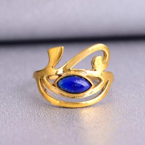 Puede incluir: Anillo dorado con una piedra azul ovalada en el centro. El anillo tiene un diseño abierto y artístico con líneas curvas. El anillo se muestra sobre un fondo neutro.