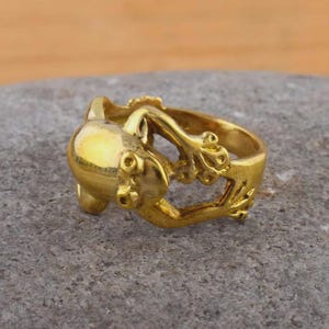 May include: A gold-colored ring featuring a stylized frog design. The ring has a rounded body and intricate details, including textured patterns and small circular accents. The frog appears to be in a crouching position.