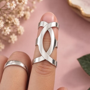 May include: A silver-toned metal finger splint ring featuring an elongated, open-work design with two intersecting bands that form a central oval shape. A simple, smooth, polished silver-toned band ring is worn on the adjacent finger.