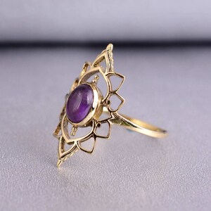 May include: A gold ring with a purple amethyst gemstone set in a delicate, intricate design. The ring features a mandala-like pattern with a central stone.