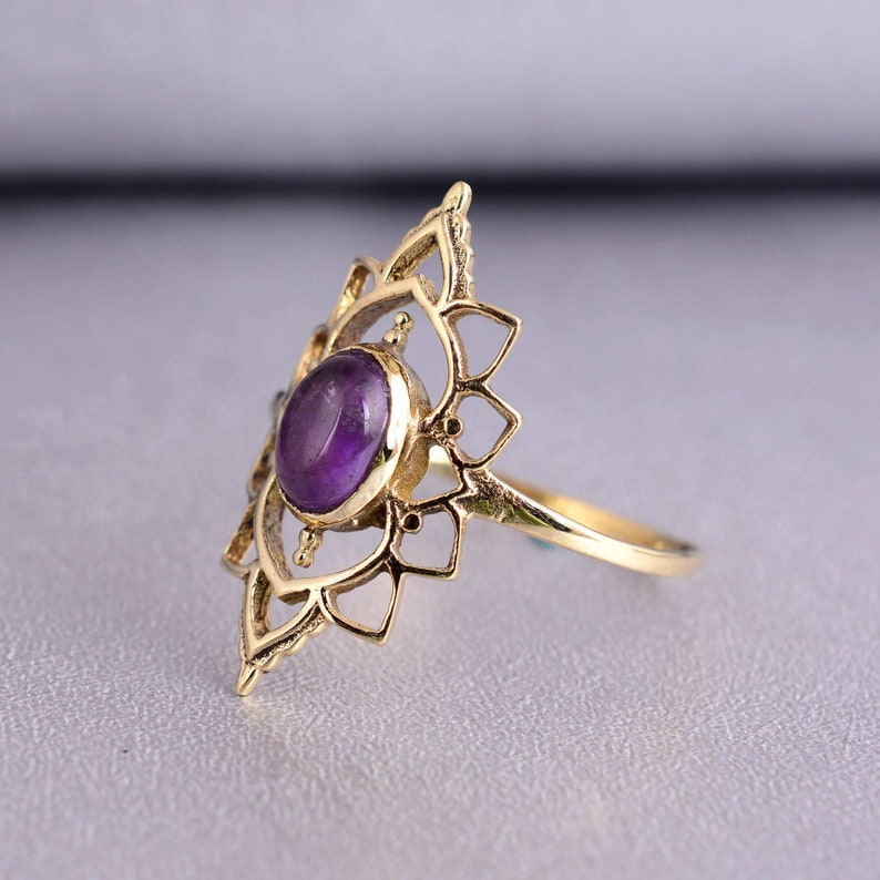 May include: A gold ring with a purple amethyst gemstone set in a delicate, intricate design. The ring features a mandala-like pattern with a central stone.