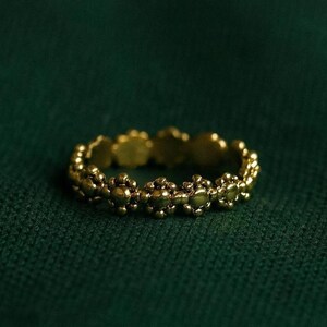 Puede incluir: Un anillo dorado con un diseño floral repetido. El anillo se muestra sobre un fondo verde oscuro, resaltando sus intrincados detalles y su cálido tono metálico. El diseño del anillo presenta pequeñas formas florales redondeadas.