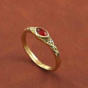 Peut inclure: Une bague en or avec une pierre rouge en forme de marquise. La monture présente des détails de filigrane complexes sur les côtés. La bague est posée sur un fond brun-rougeâtre chaud.