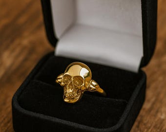 May include: A gold skull ring presented in a black velvet ring box. The ring has a detailed skull design, including eye sockets, a nasal cavity, and teeth. The ring is set against a white interior and a wooden surface.