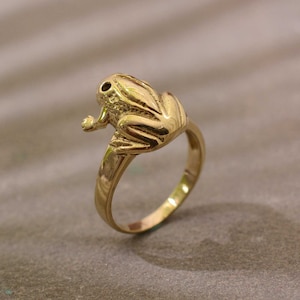 Könnte beinhalten: Ein goldener Froschring. Der Frosch sitzt auf dem Ringband.
