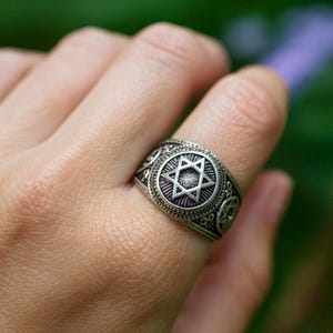 Può includere: Anello color argento con un design a Stella di David. L'anello presenta un motivo dettagliato attorno alla fascia e una stella a sei punte prominente al centro. L'anello è indossato su un dito.
