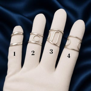 May include: Four silver rings of varying designs are displayed on fingers covered in a white glove. The rings include a simple band, a ring with a "C" shape, a ring with a "U" shape, and a ring with two crescent shapes. Each ring is labeled with a number.