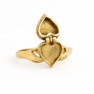 Op de afbeelding: Een goudkleurige ring met een hartvormig medaillon dat opengaat. De ring heeft een hartvormig ontwerp met een scharnierende bovenkant, waardoor een holle binnenkant zichtbaar wordt. De band van de ring heeft aan weerszijden van het hart een handachtig ontwerp.