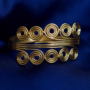 May include: Gold-toned arm cuff with a spiral design. The cuff features multiple concentric circles and curved lines, creating an elegant and eye-catching accessory. The cuff is displayed against a dark blue background.