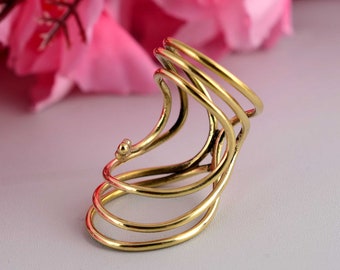 May include: A gold-colored ear cuff with a unique, swirling design. The cuff features multiple curved bands that wrap around the ear, creating an elegant and modern look. The jewelry is set against a soft pink and white background.