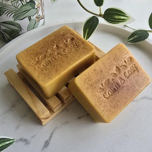 May include: Two rectangular, light brown soaps with the embossed text "Calm & Cozy" rest on a small wooden soap dish. The soaps sit on a white, round tray with decorative greenery and a patterned planter in the background.