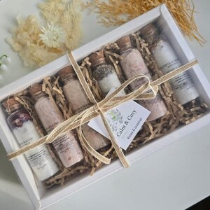 May include: A white box with a clear lid contains several small glass bottles filled with bath salts. The bottles are arranged on shredded paper and tied with twine. A small tag reads "Calm & Cosy, Relax & unwind."