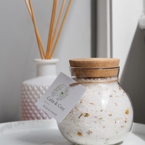 May include: A round glass jar filled with white bath salts, sealed with a cork lid. A label reads "Calm & Cosy" with a botanical design. The jar is on a white tray, with a white vase and reeds in the background.