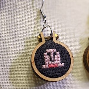 May include: A pair of wooden hoop earrings with a black fabric center. One earring features a pink and white cross-stitch design of a ghost wearing a bow tie.