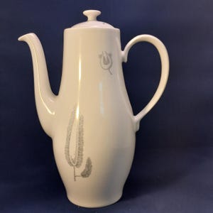 Può includere: Una caffettiera alta in ceramica bianca dotata di un beccuccio sottile, un manico curvo e un coperchio abbinato. Il corpo della caffettiera è decorato con delicati disegni a linee grigio chiaro raffiguranti motivi botanici.