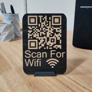 May include: A black wooden sign featuring a QR code and the text "Scan For Wifi". The sign has rounded corners and is supported by a small, dark-coloured stand. The QR code and text are light-coloured.