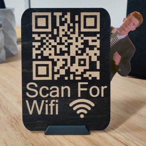 Może przedstawiać: Czarny drewniany znak z kodem QR i białym napisem "Scan For Wifi". Znak ma małą, ciemnoniebieską podstawę. Wycięcie mężczyzny w garniturze wystaje zza znaku.