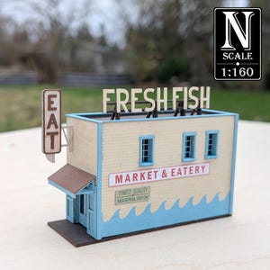 May include: A miniature model of a fish market and eatery. The building has a beige exterior with blue trim and a wave design. The sign reads "FRESH FISH" and "MARKET & EATERY". The scale is 1:160.