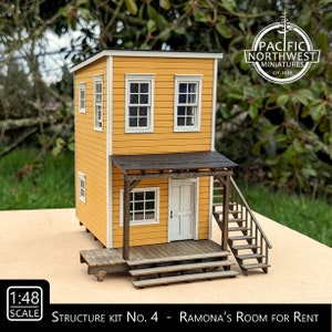 May include: A 1:48 scale model kit of a two-story building with a porch and stairs. The building is yellow with white trim and has a brown roof. The kit is called "Ramona's Room for Rent" and is made by Pacific Northwest Miniatures.