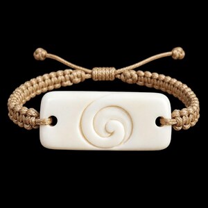 May include: A bracelet featuring a rectangular, off-white, smooth center piece with a carved spiral design. The center piece is attached to a tan, braided cord band that includes an adjustable sliding knot closure with two small knotted ends.