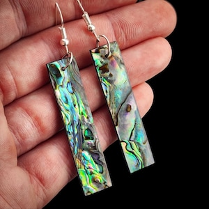 Può includere: Un paio di orecchini pendenti rettangolari con inserti in conchiglia di abalone iridescente caratterizzati da vivaci vortici di verde, blu e viola. Gli orecchini sono fissati a ganci in metallo color argento e presentano una finitura lucida.