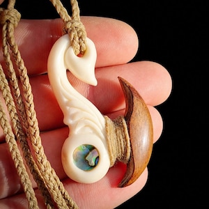 May include: A pendant featuring a cream-colored carved hook shape with an iridescent abalone shell inlay and a dark brown wooden accent piece, all secured with tan braided cord wrapping and suspended from a matching braided cord necklace.