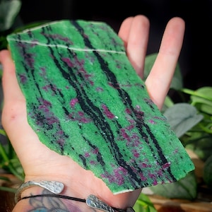 May include: A green and red zoisite slab with black streaks. The natural stone has an irregular shape and a polished surface. The colours are a mix of deep green, ruby red, and black, creating a striking contrast. The stone is a unique decorative item, perfect for display.