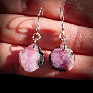 May include: A pair of drop earrings featuring raw, irregular pink tourmaline stones with black inclusions. Each stone is suspended from a silver-toned metal hook by a circular jump ring, showcasing a natural, unpolished texture and vibrant pink hues.
